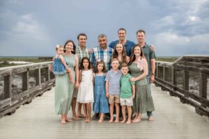 Large multi-generational family in coordinating sage green, blue & denim tones posing on a beach boardwalk in Mexico Beach, Florida for an extended family portrait session